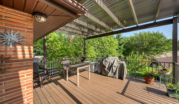 Custom deck on a brick home. Covered grill, patio furniture, and potted plants on the deck. View of trees in background.