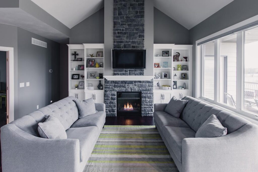 Gray living room with fireplace, vaulted ceiling, and row of large windows on right side.