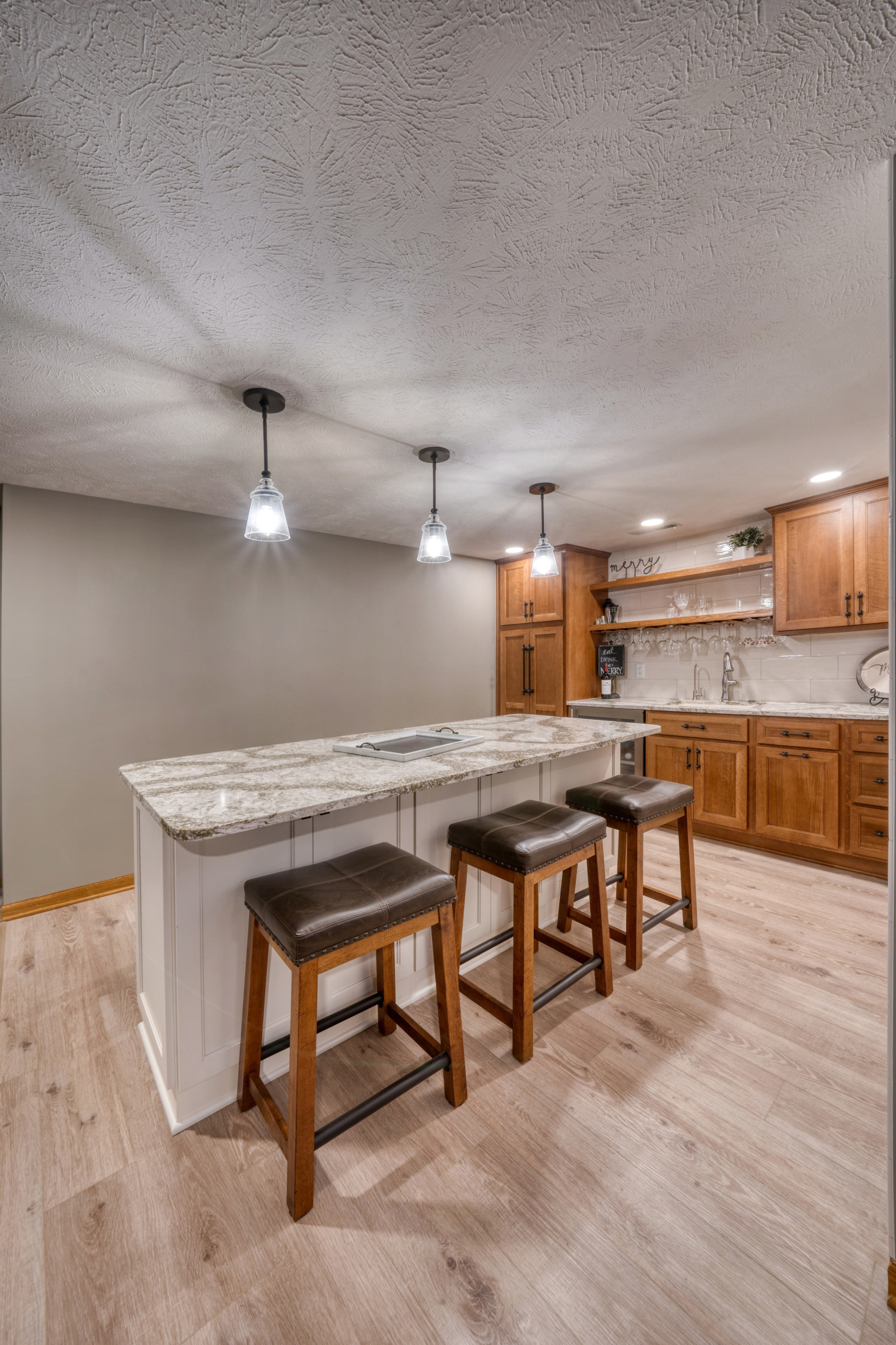 Remodeled basement kitchen with recessed lighting, a large island with pendant lighting overhead, and wooden cabinetry with a white backsplash.