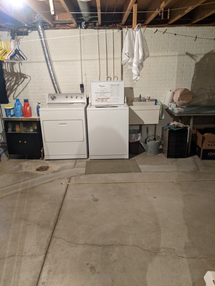 White washer and dryer in an unfinished basement with cement floor and exposed beams and pipes in ceiling.