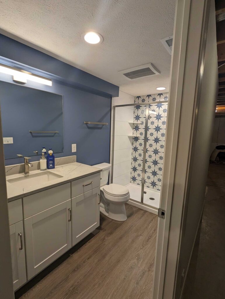 New basement bathroom with light wood floors, gray-blue wall, white vanity with stone countertop, blue patterned tile on shower wall, and recessed lighting.