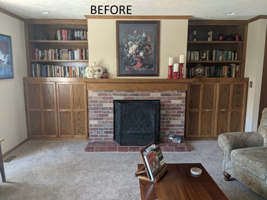 "Before" image of brick fireplace and outdated woodgrain built-in shelving and cabinets on either side, in tan-carpeted living room.