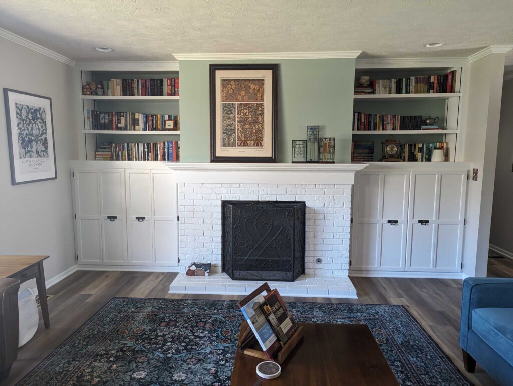 "After image of white-pained brick fireplace and white built-in shelving and cabinets on either side, in living room with pale green accent wall above fireplace and new wood flooring.