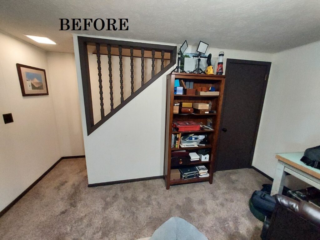 Outdated Before picture of carpeted basement area with wall and outdated railing along stairs.