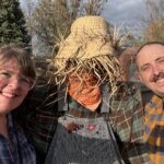 A couple of people smiling around a scarecrow